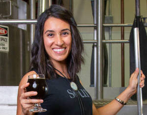 Ayla Kapahi smiling while holding a craft brew, standing in a distillery with brewing equipment in the background.