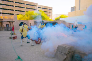 Men in historical military clothing firing a replica cannon at the Presidio Museum with embers and smoke, Downtown Tucson skyscrapers visible in the background.
