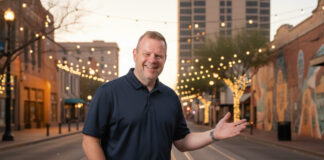 Tom Heath standing in Downtown Tucson at dusk, gesturing warmly amid string lights and city landmarks like the Fox Theatre and streetcar tracks, capturing the vibrant energy of Tucson’s November festivals.